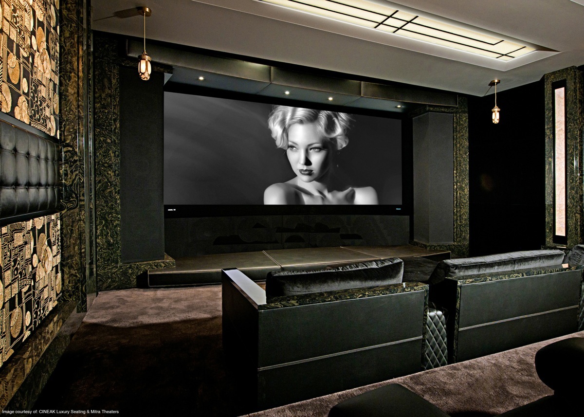 Luxurious home theater with tiered rows of plush black seating facing a large projection screen displaying a black-and-white portrait of a woman, surrounded by dark patterned walls and pendant lights.