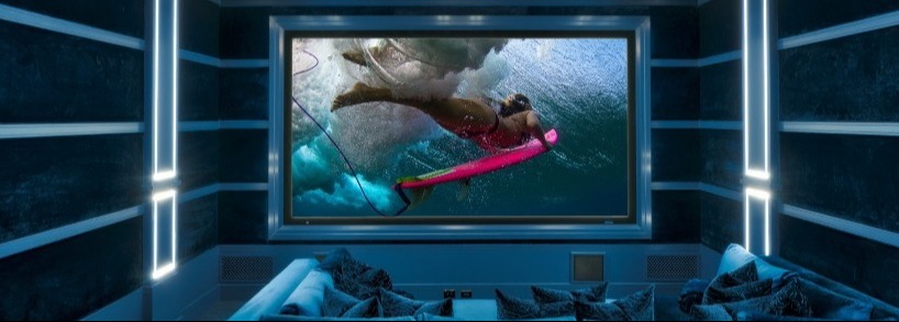 <span class="uk-h4">Home theater with a large screen showing an underwater surfing scene, viewed from a cozy front row of blue cushioned seating and pillows.</span>