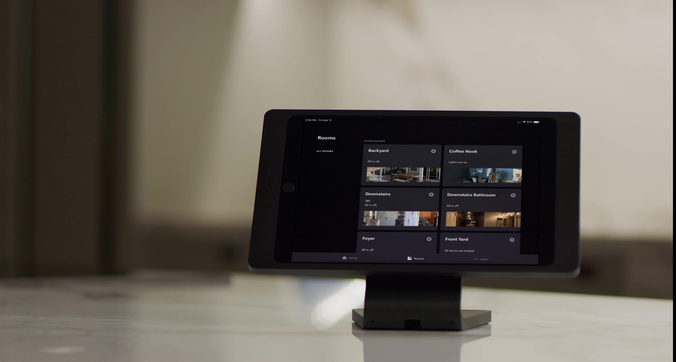 Tablet on a stand displaying a smart-home dashboard with tiles for rooms like Backyard, Coffee Nook, Downstairs, Foyer, and Front Yard—status shows lights and locks—sitting on a marble counter with a softly blurred interior in the background.