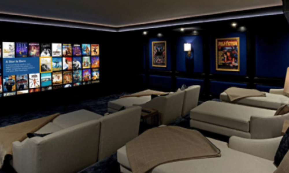 <span class="uk-h4">Cozy home theater with several light-colored reclining seats facing a wall-mounted screen of movie covers, dark blue walls, and framed posters.</span>